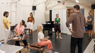Seven adults are chatting and smiling in a relaxed, informal indoor space with white walls and fairy lights. Some are sitting and others are standing, suggesting a casual gathering or rehearsal. A piano and chairs are visible in the background.