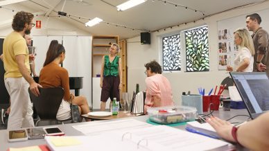 A group of people gathers in a bright, modern office or studio, engaged in discussion. Papers, laptops, and stationery are spread on a large table in the foreground, with windows and posters on the wall behind them.