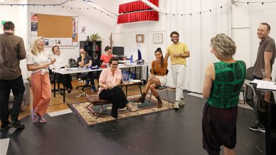 A group of people in a brightly lit, casual office space are laughing and talking. Some are seated at desks with computers, whilst others stand or sit nearby, creating a relaxed and friendly atmosphere.
