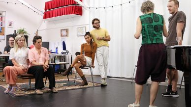 Six adults participate in a casual indoor meeting or rehearsal. Four sit attentively, two stand, and one stands with their back to the camera, speaking. The group is in a creative studio with eclectic decor and fairy lights.