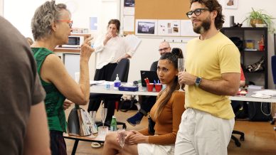 A group of people interact in an office; a woman gestures while talking to a man holding a drink, as others sit or work at desks in the background. The environment is casual and relaxed.