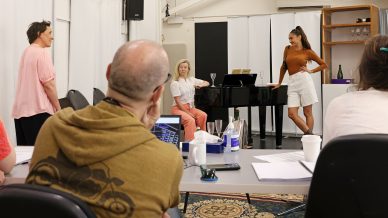 Four people are rehearsing in a music or theatre studio; two women stand and talk near a piano, whilst another woman sits at the piano and a man in a yellow hoodie observes from a table covered with papers.