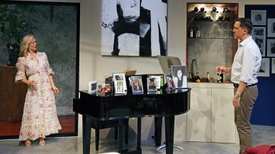A woman in a floral dress and a man holding a glass stand in a modern room by a black piano covered with framed photos, with art on the wall and shelves with bottles in the background.
