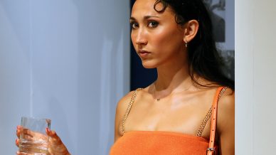 A woman in an orange dress with gold chain straps stands by a wall, holding a glass of water. She has long hair pulled back, wears hoop earrings, and carries an orange handbag on her shoulder.