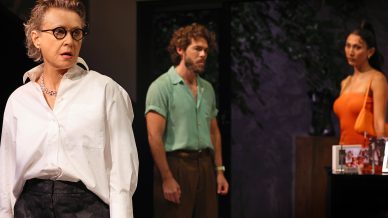 A woman in glasses and a white shirt stands in the foreground, looking surprised. Behind her, a man in a green shirt and a woman in an orange dress stand facing each other in a dimly lit room.