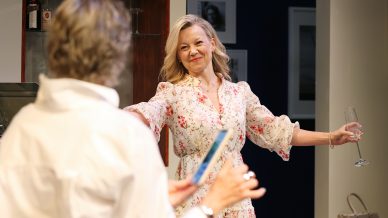 A woman in a floral dress smiles warmly with her arms open, holding a glass, whilst another person in a white shirt faces her, holding a blue object. They appear to be in a warmly lit room, possibly at a social gathering.