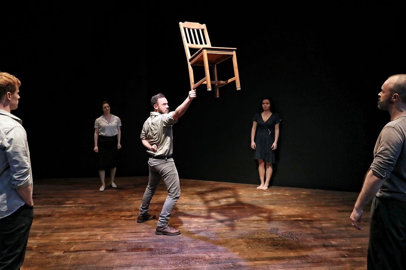 A man balances a wooden chair on one hand in the centre of a wooden stage, surrounded by four people standing and watching against a dark background.
