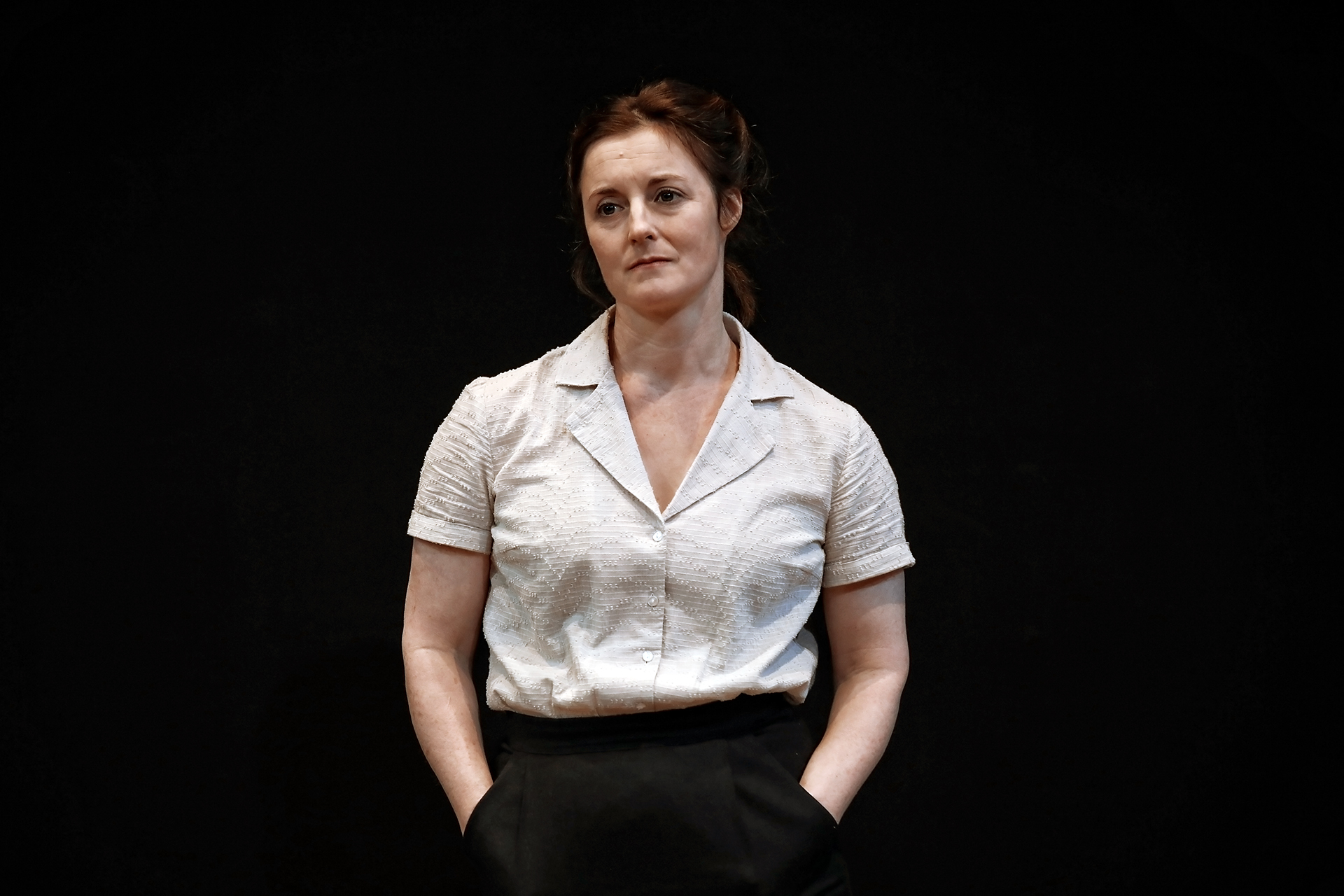 A woman stands against a dark background, wearing a short-sleeved, light-coloured blouse and dark trousers, with her hands in her pockets and a neutral expression on her face.