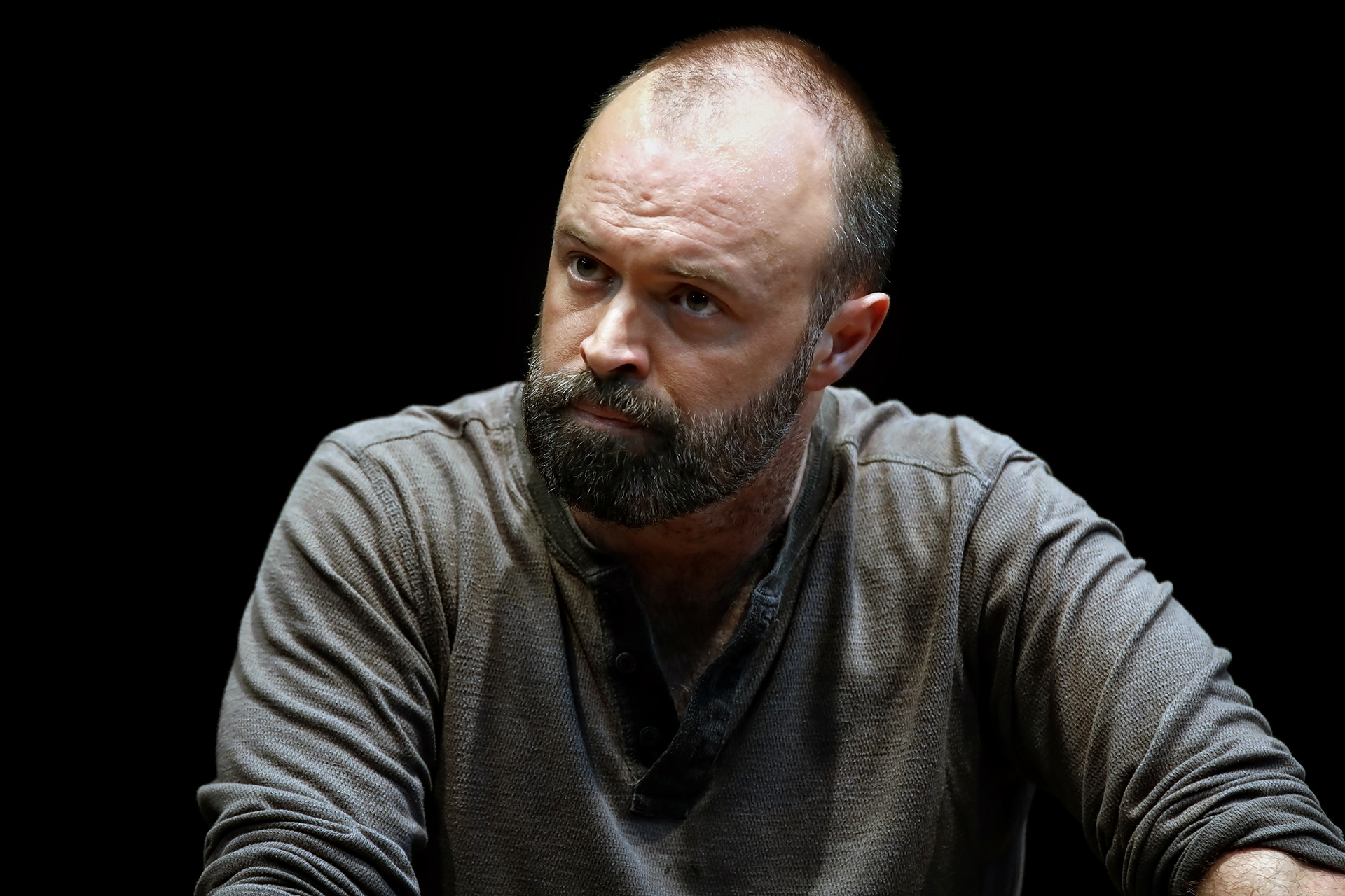 A man with a beard and receding hairline, wearing a grey buttoned shirt, sits against a dark background, looking slightly to the side with a serious expression.