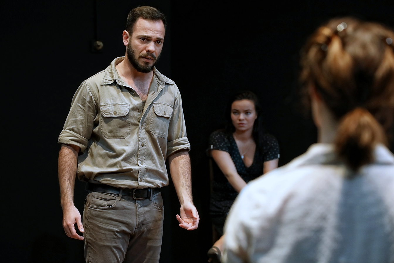 A man in a light grey shirt and trousers stands facing a woman with her back to the camera, while another woman sits in the background, watching them intently in a dark room.