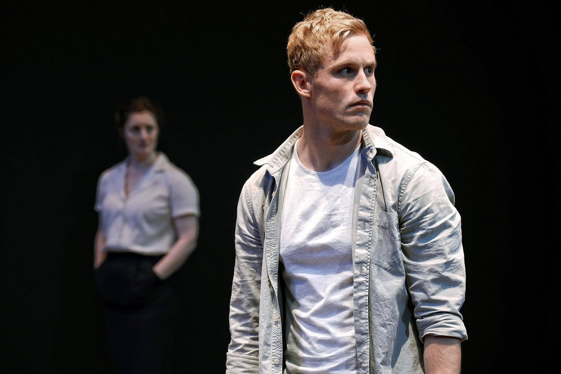 A man in a light shirt stands in the foreground, looking to the side with a serious expression, whilst a woman in a blouse and dark skirt stands blurred in the background against a dark backdrop.
