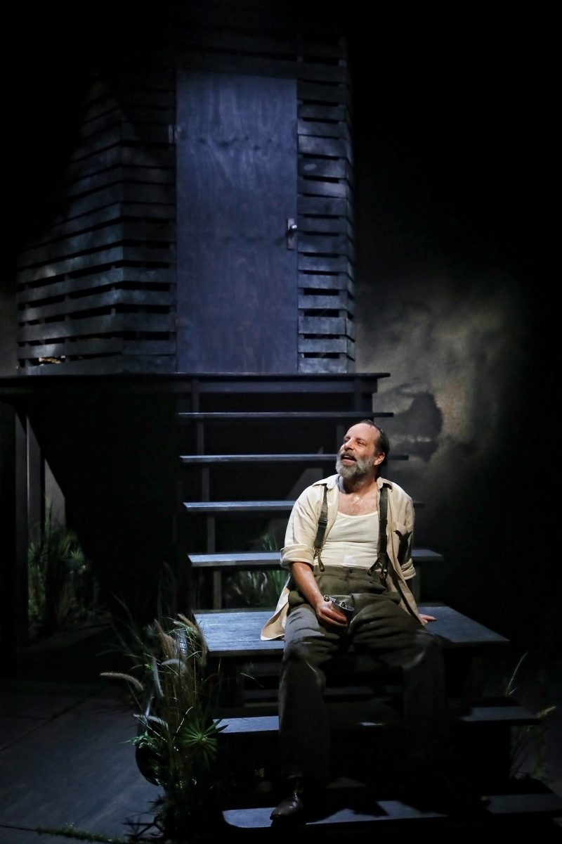A bearded man in braces sits on steps in front of a dark wooden structure on a dimly lit stage, holding a small object and looking upwards thoughtfully.