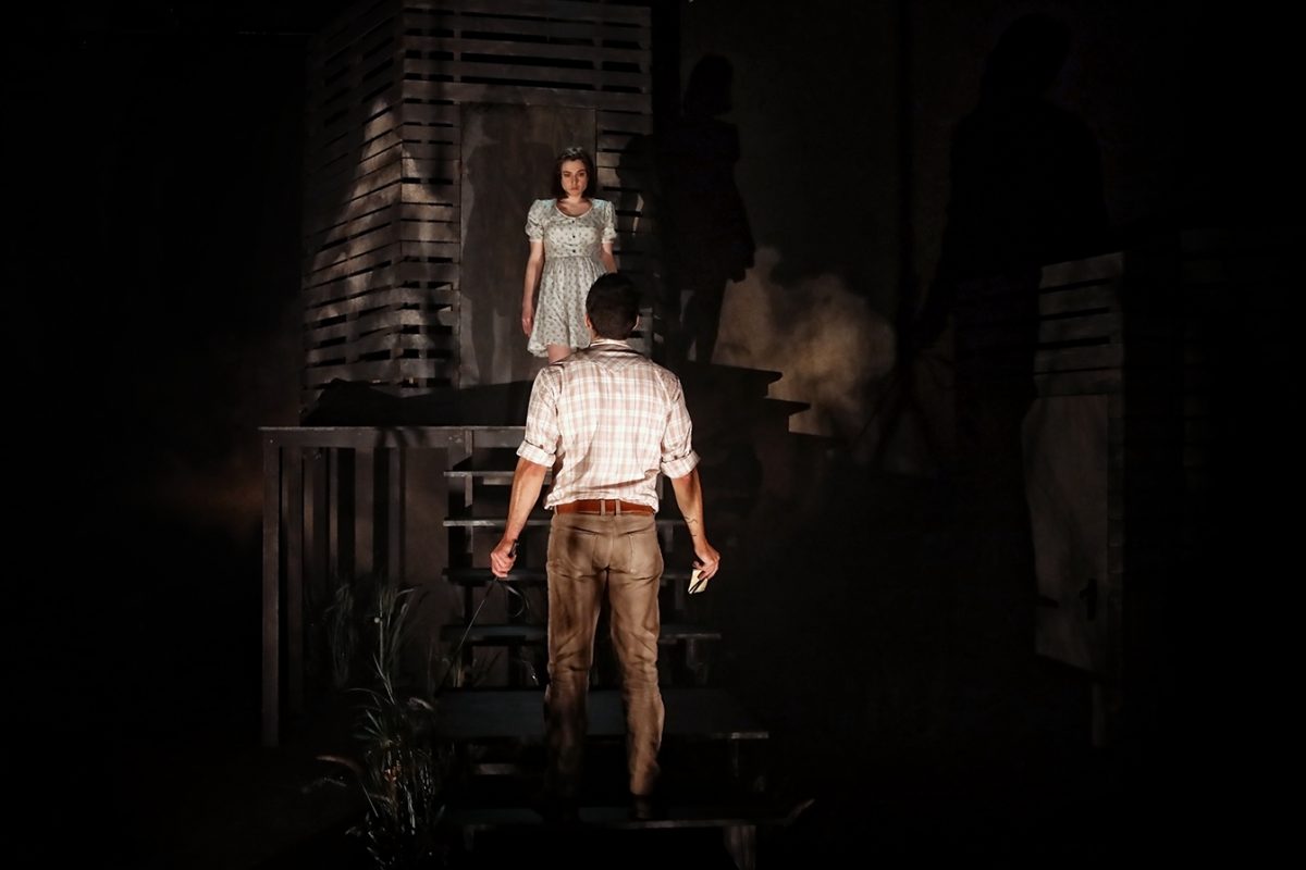 A man in a checked shirt stands at the bottom of the stairs, facing a woman in a light dress who stands above him on a platform in a dimly lit, dramatic stage setting with wooden structures and shadows.