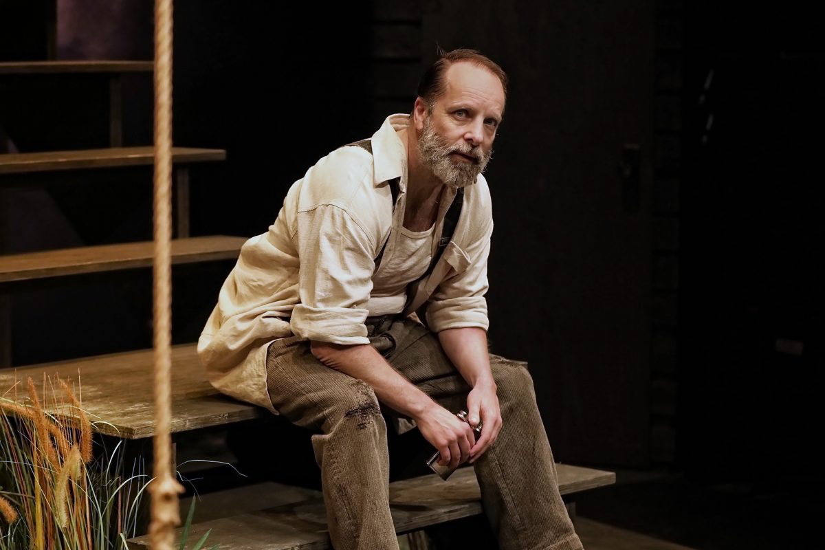 A middle-aged man with a beard, wearing a light shirt and brown trousers, sits on wooden steps indoors. He looks thoughtful, with hands clasped and elbows resting on his knees. Some tall grass is visible in the foreground.