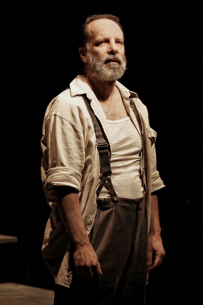 A middle-aged man with a beard stands in dramatic lighting, wearing a loose shirt, braces, and a white vest, looking upward with a serious expression.