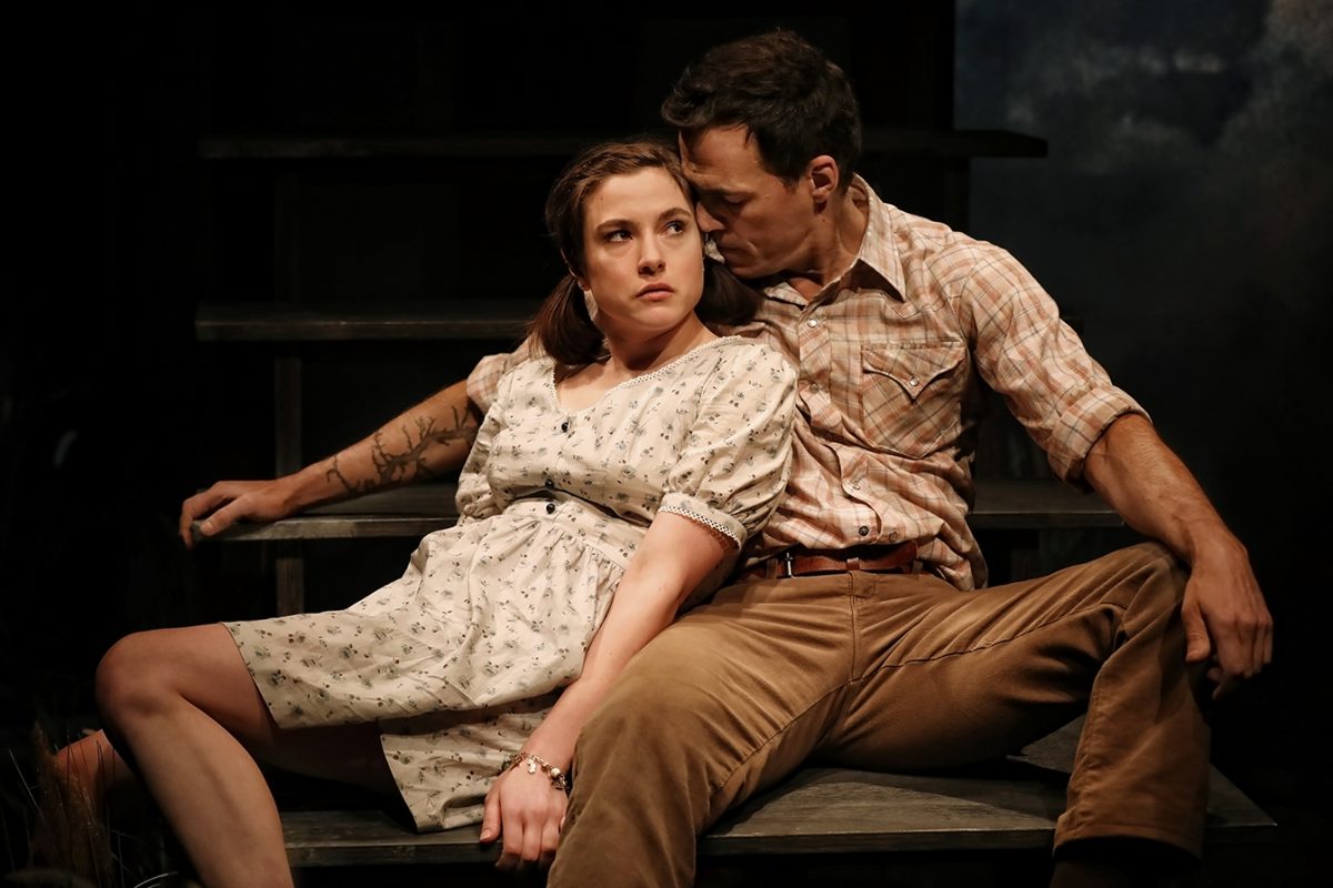 A woman in a light dress sits on wooden steps beside a man in a checked shirt and brown trousers. She looks ahead with a serious expression whilst he leans in close, appearing to whisper or speak softly to her.