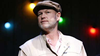 A man with a trimmed beard wears a flat cap and light jacket, looking slightly upwards. Colourful string lights in yellow, red, blue, and green are blurred in the background.