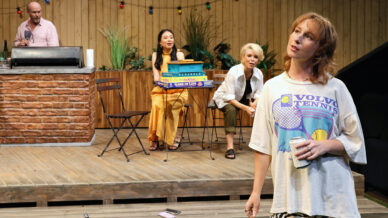 A woman in a tennis T-shirt stands in the foreground holding a drink, whilst three people sit behind her on a wooden patio. One holds a stack of board games. Fairy lights and plants decorate the background.