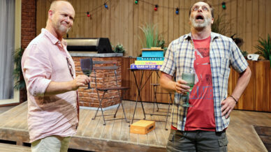Two men stand on a wooden terrace holding wine glasses; one looks amused, the other looks up with his mouth open. Board games are stacked on a table behind them, and fairy lights and plants decorate the background.