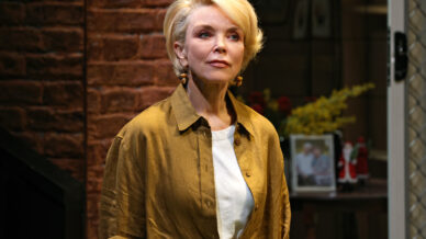 A woman with short blonde hair, wearing a mustard brown button-up over a white shirt, stands indoors in front of a brick wall and a shelf with framed photos and flowers. She looks thoughtful.