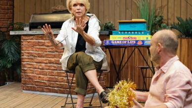 A woman sits on a chair gesturing with her hands, while a man sits on the floor holding gold tinsel. Behind them is a stack of board games on a small table and a barbecue on a wooden decking with plants.