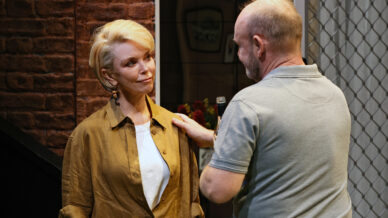 A woman with short blonde hair and a tan shirt stands as a man in a grey polo gently touches her shoulder and holds a microphone, both appearing engaged in a serious conversation on stage.