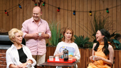 Four adults sit and stand around a small outdoor table with coffee cups. One man wears novelty reindeer antlers and smiles, while the three women laugh and chat. Colourful fairy lights and plants decorate the wooden fence behind them.