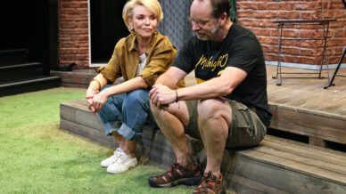 A woman and a man sit on wooden steps outside, with a lawn in front of them. The woman, wearing a brown shirt and jeans, looks at the man, who wears a black T-shirt and shorts. They both appear thoughtful.
