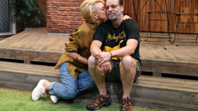 A woman kisses a man on the cheek as they sit on wooden steps outdoors. She wears a brown shirt, jeans, and white trainers; he wears glasses, a black t-shirt, shorts, and sandals. There are chairs and a table in the background.