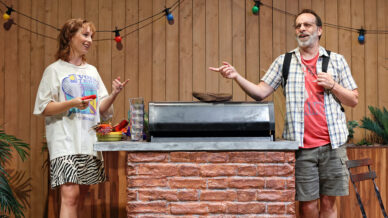 Two people stand by an outdoor barbecue bar decorated with fairy lights. The woman, wearing a graphic T-shirt and zebra-print skirt, smiles while holding a drink. The man, in casual shorts and a rucksack, gestures as they converse.