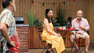 Three people are on a wooden patio set. A woman in an orange dress sits at a small table with a surprised expression, whilst a man in light clothes gestures beside her. Another man stands in the foreground, facing them.