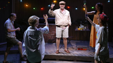 Five people stand on a patio stage raising glasses in a toast, with fairy lights overhead and drinks on a table. The person in the centre wears a hat and beige outfit, while others are casually dressed. The atmosphere is festive.