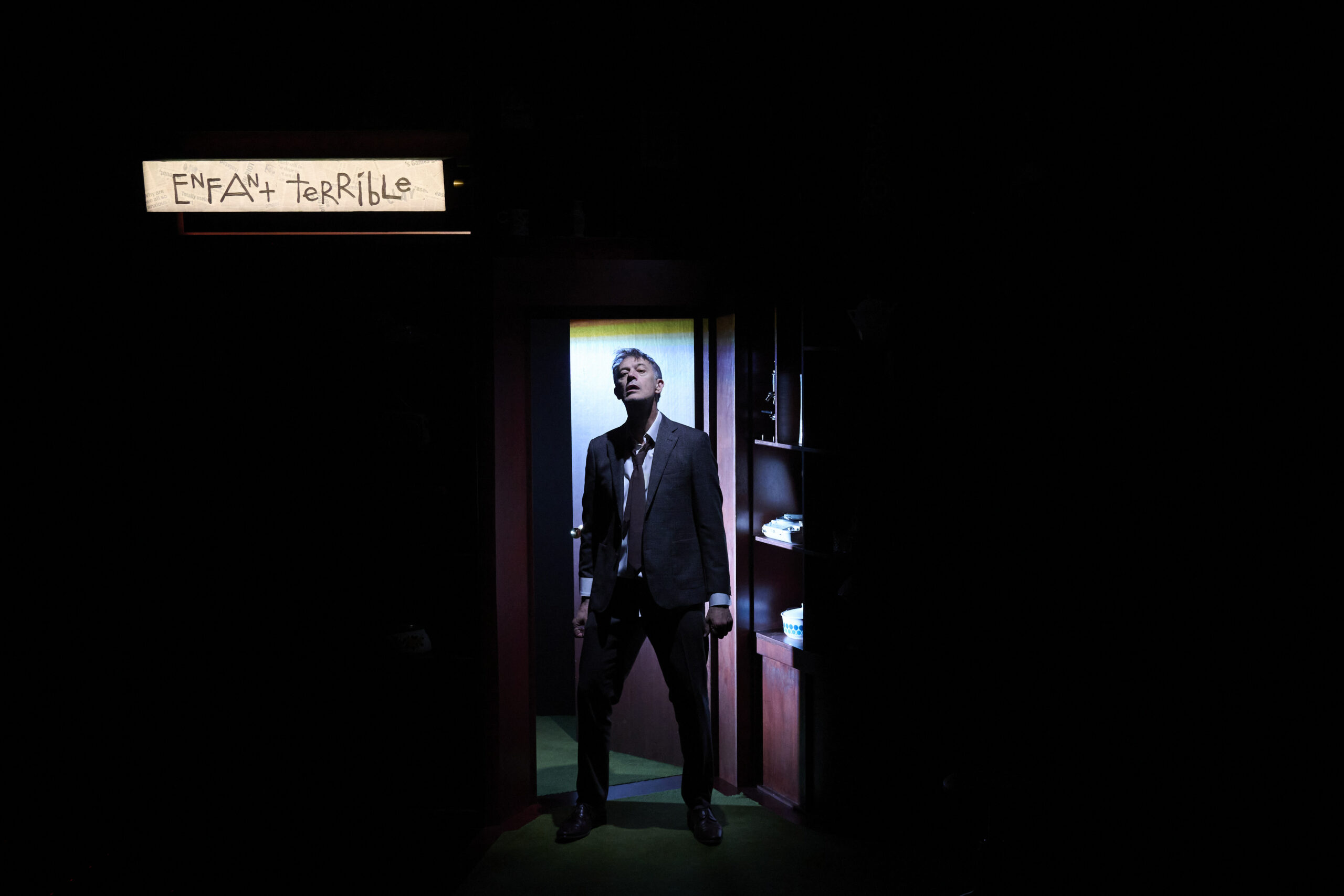 A man in a suit stands dramatically in a doorway, lit by a spotlight, with a sign above reading ENFANT TERRIBLE in handwritten letters. The surrounding area is mostly dark.