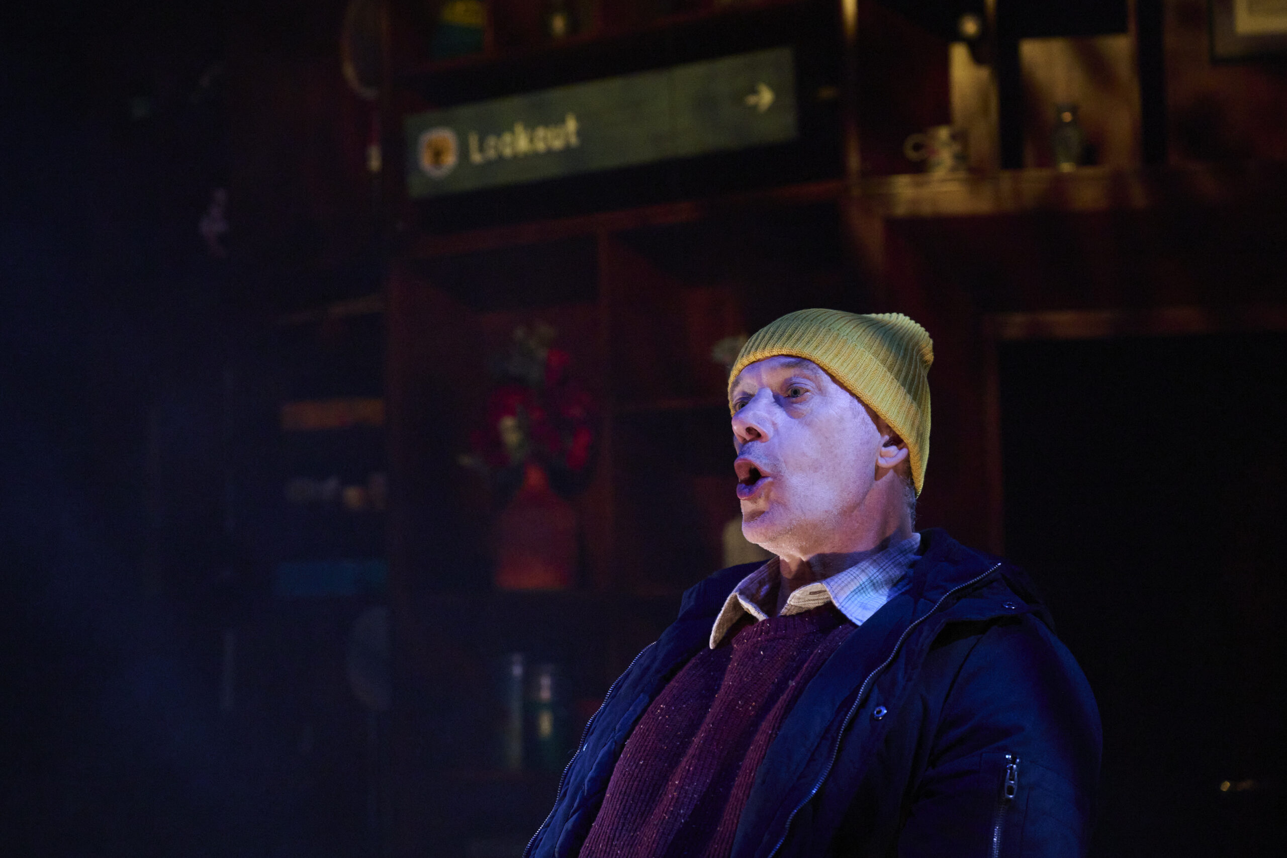 A man wearing a yellow beanie and dark jacket appears to be speaking or singing with an expressive face. Behind him, shelves and a sign that reads Lookout are visible in a dimly lit setting.