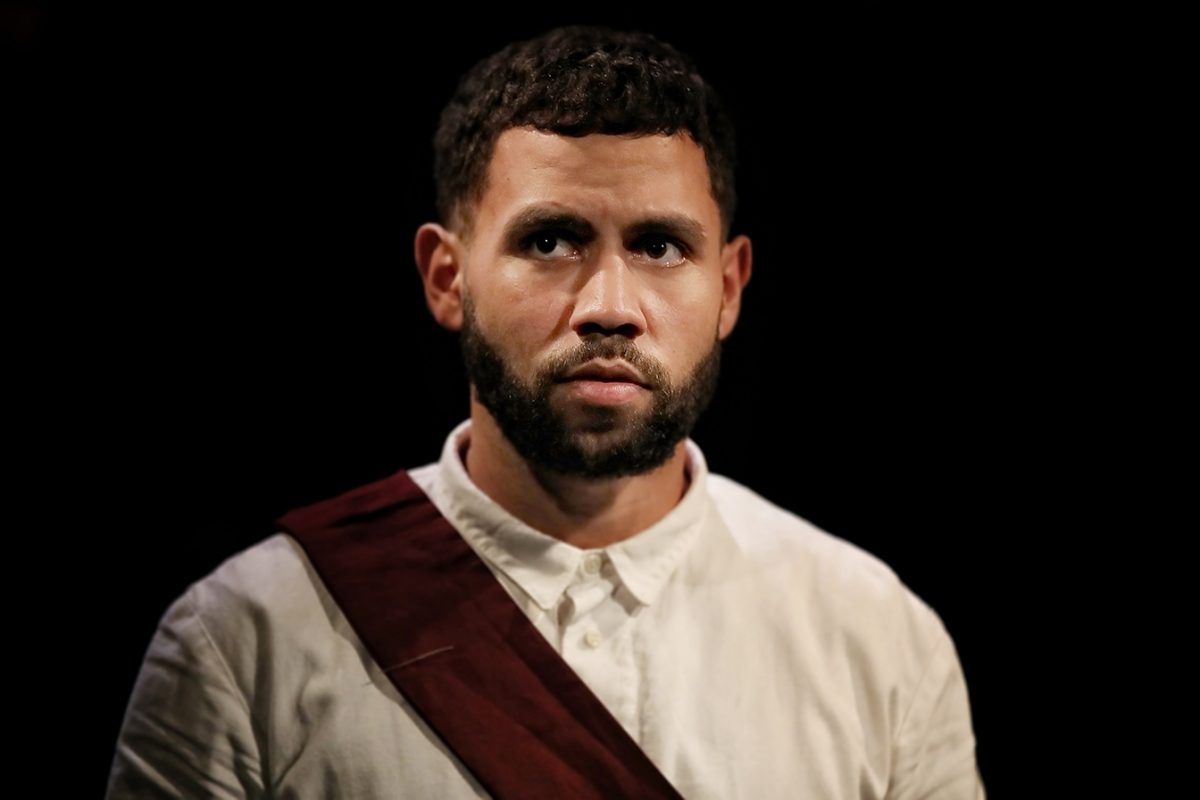 A man with short curly hair and a beard wears a white shirt and a maroon sash over one shoulder, standing against a black background and looking slightly to the side with a serious expression.