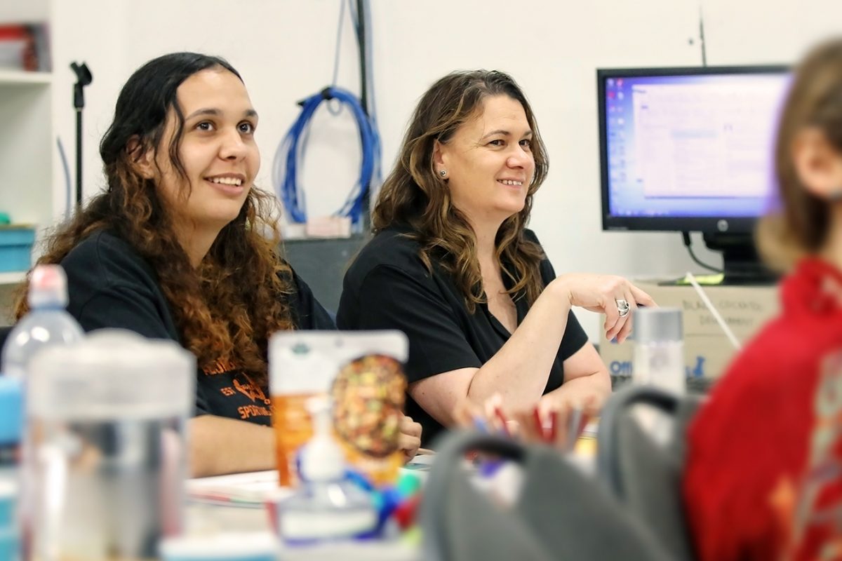 Two women sitting at a table, smiling and looking ahead. The table is scattered with various items and there is a computer monitor in the background. The environment looks like a classroom or office setting.