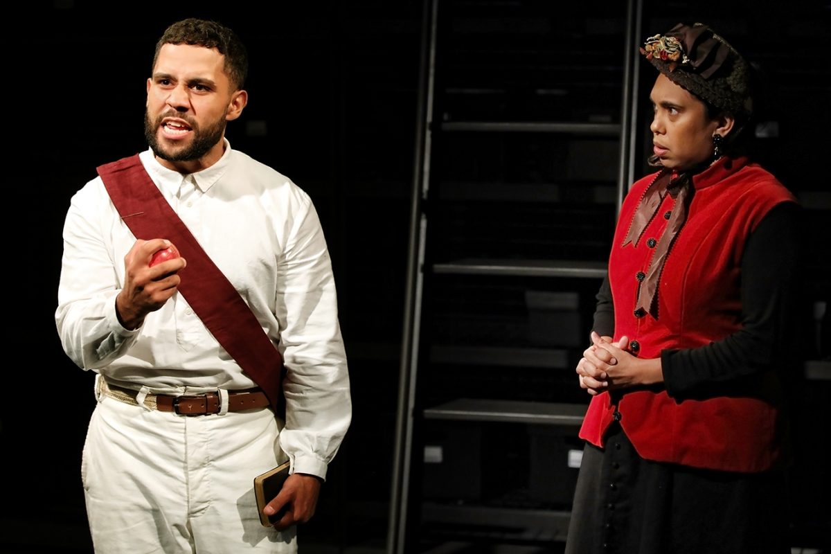 Two actors perform on stage. The person on the left, dressed in white with a maroon sash, gestures passionately while holding an object. The person on the right, in a red and black outfit, stands watching with hands clasped.