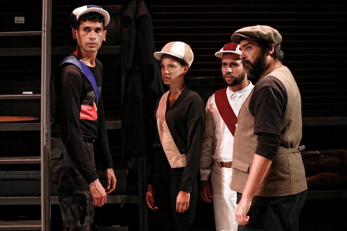 Four people wearing costumes with caps and sashes stand together indoors, looking in the same direction with serious expressions. Shelves and dim lighting suggest a backstage or theatrical setting.