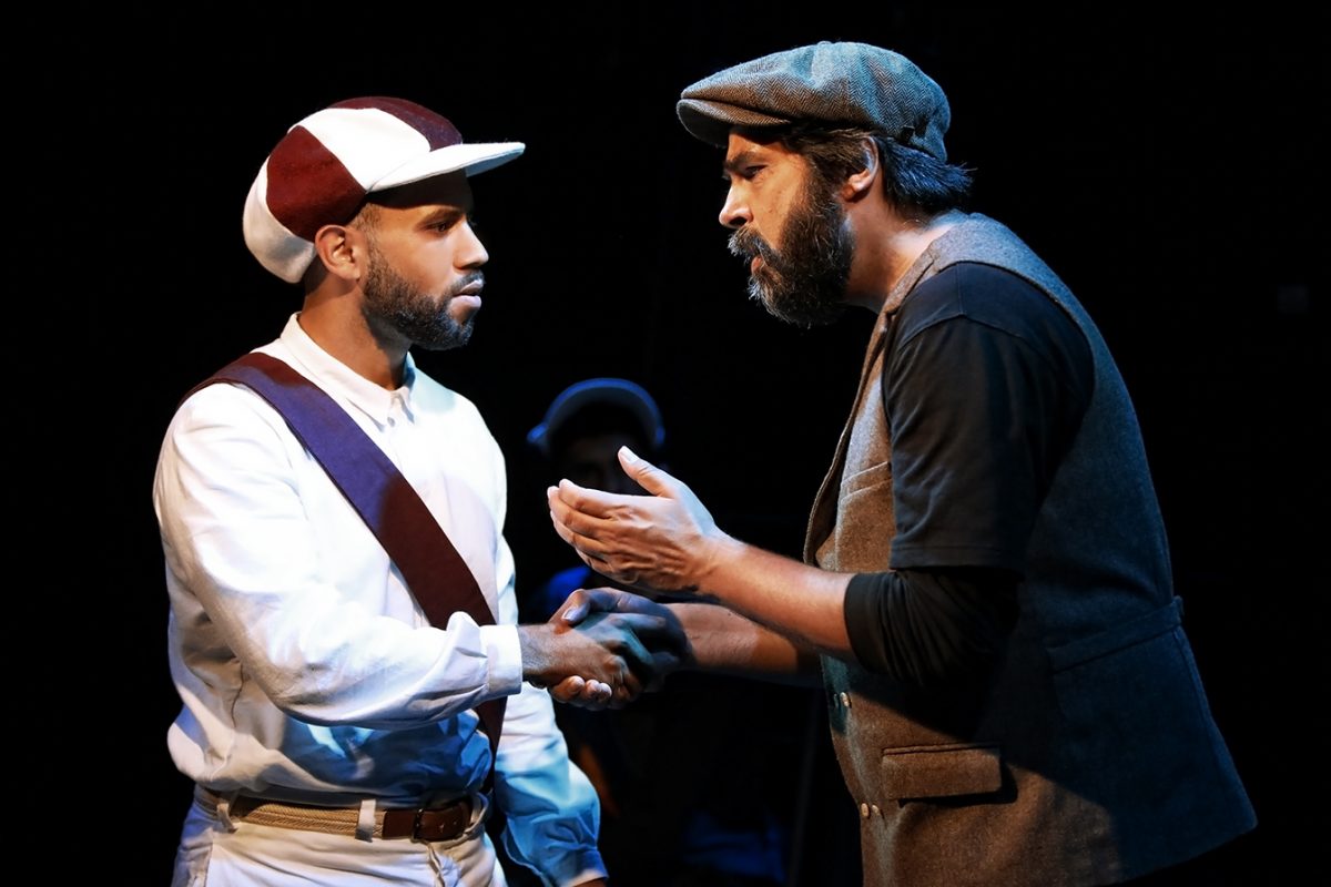 Two men are shaking hands on stage; one wears a vintage baseball kit and cap, while the other wears a flat cap and waistcoat, gesturing earnestly as they converse under dramatic lighting.