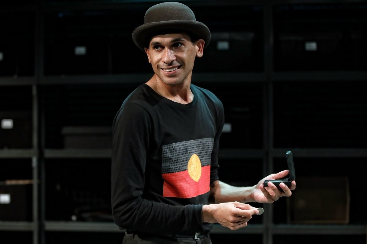 A man wearing a black bowler hat and a black shirt with the Aboriginal flag design stands indoors, smiling and holding a small black box, against a background of dark shelves.
