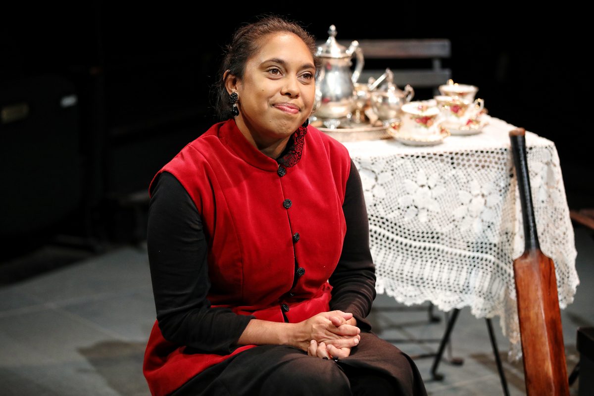 A woman in a red waistcoat sits with hands clasped, smiling slightly. Behind her is a table with a lace cloth, tea set, and teapot, along with a wooden bat leaning on the table. The setting appears to be a stage performance.
