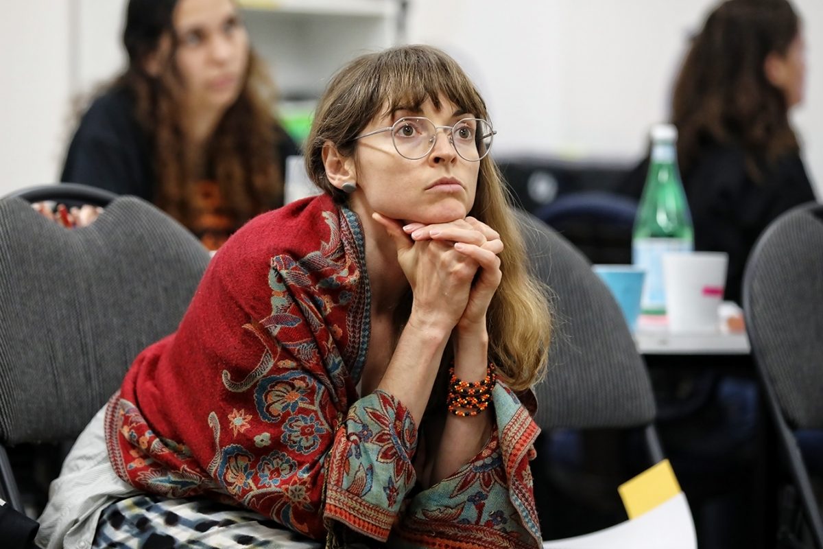 A woman with glasses and long hair, wrapped in a red patterned shawl, sits with her chin resting on her clasped hands, looking thoughtful in a classroom or meeting setting. Other people are blurred in the background.
