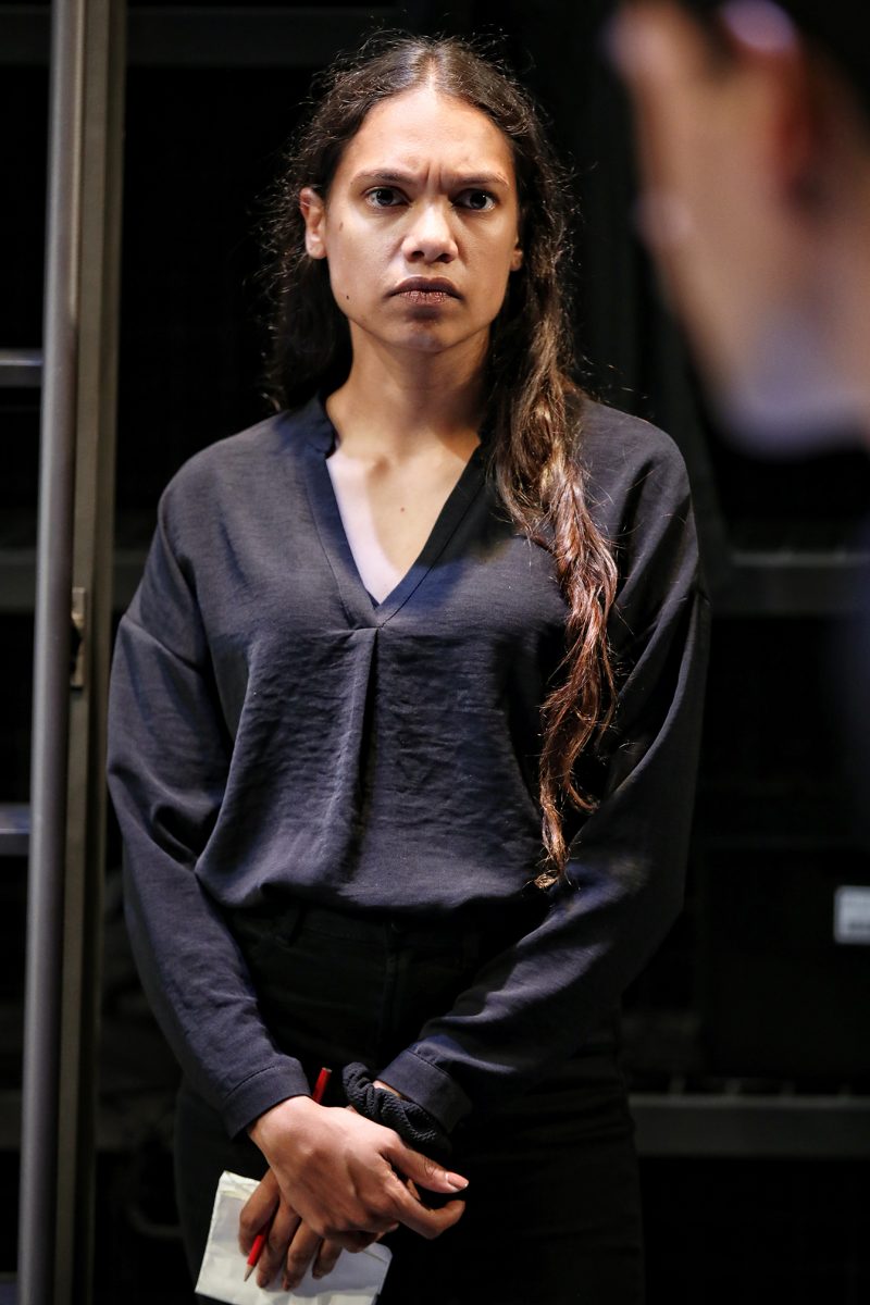 A person with long, wavy brown hair stands indoors, wearing a dark V-neck shirt and black trousers, holding a sheet of paper. They have a serious expression and are looking slightly to the side.