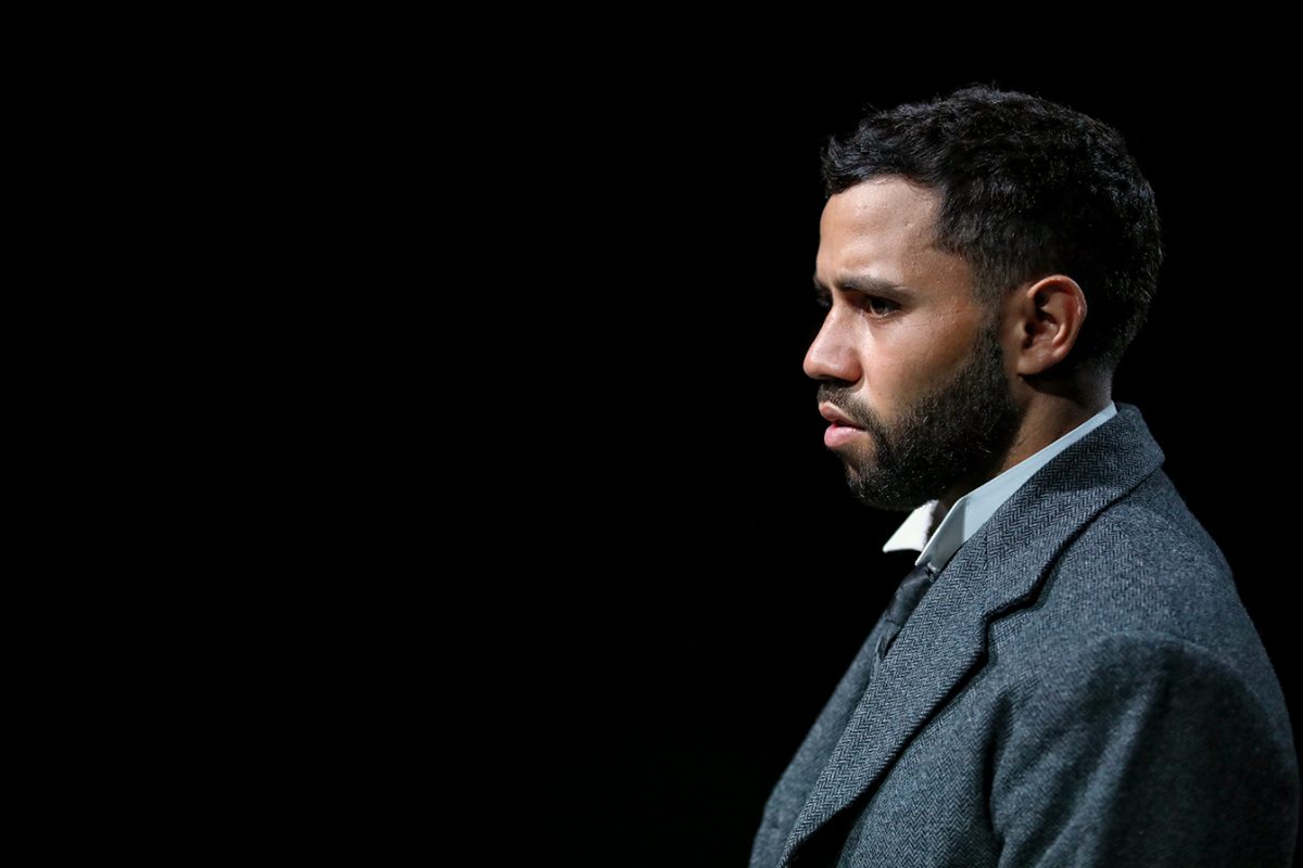 A man with short dark hair and a beard, wearing a grey suit and white shirt, stands in profile against a black background, looking serious and focused.