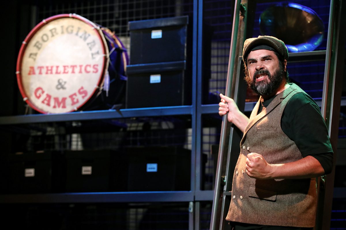 A man with a beard and wearing a cap and waistcoat stands on stage, gripping a ladder and gesturing passionately. Behind him are black storage boxes and a large drum labelled “Aboriginal Athletics & Games.”.