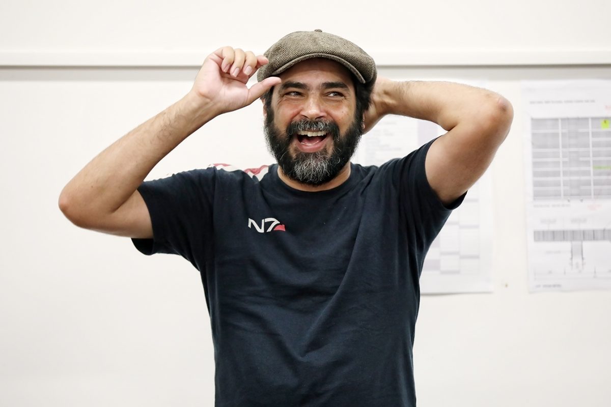 A bearded man in a black T-shirt and flat cap smiles broadly while holding his hat with one hand and touching the back of his head with the other, standing indoors in front of a white wall with papers pinned to it.