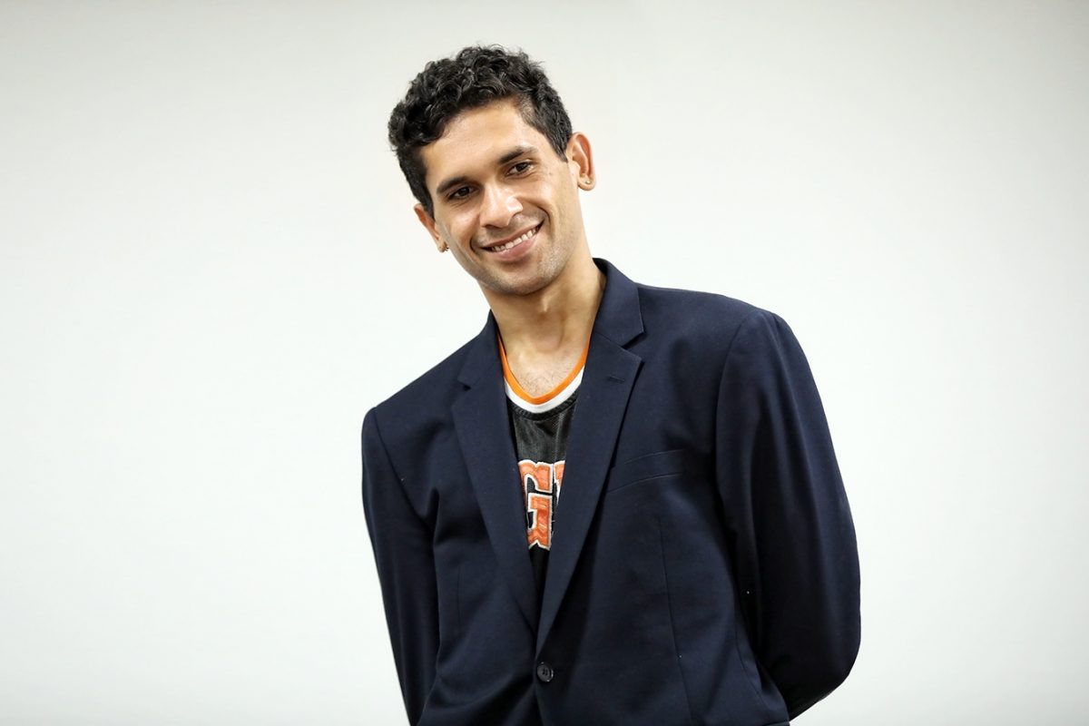 A young man with short curly hair smiles whilst wearing a dark blazer over a basketball vest, standing against a plain white background.