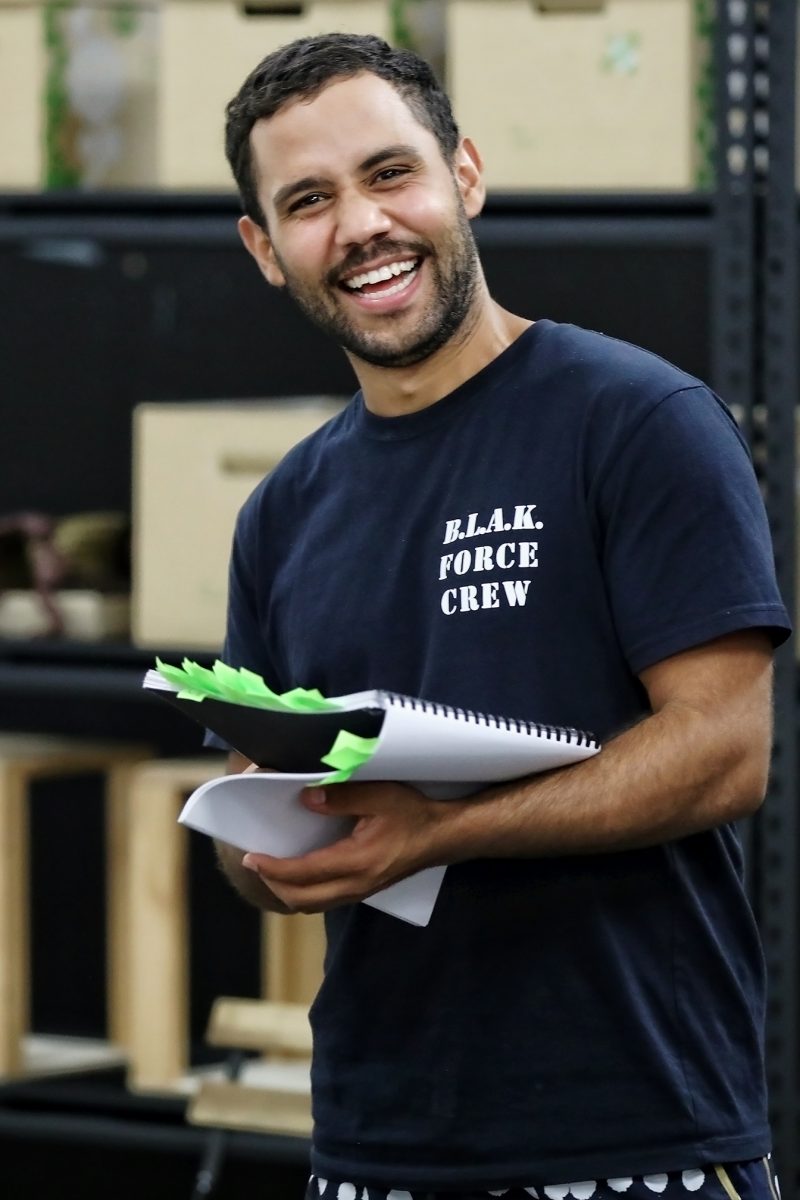 A person smiling whilst holding a spiral-bound notebook with green page markers, wearing a navy shirt that says B.L.A.K. FORCE CREW, standing indoors in front of shelves with boxes.