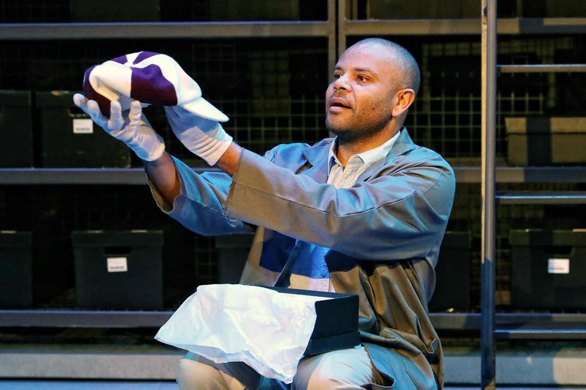A man wearing white gloves and a grey coat kneels while holding up a folded cloth with a purple and white pattern, near an open black box lined with tissue paper. Shelves with stacked black boxes are in the background.
