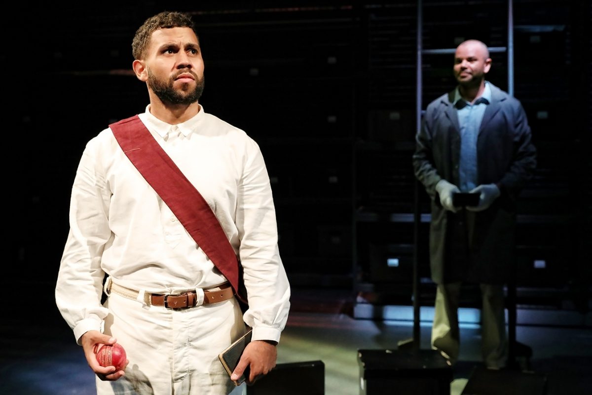 A man in white clothes with a red sash holds an apple and a book, looking ahead seriously. Another man in a coat stands in the shadowy background, holding a walking stick, both on a dimly lit stage.